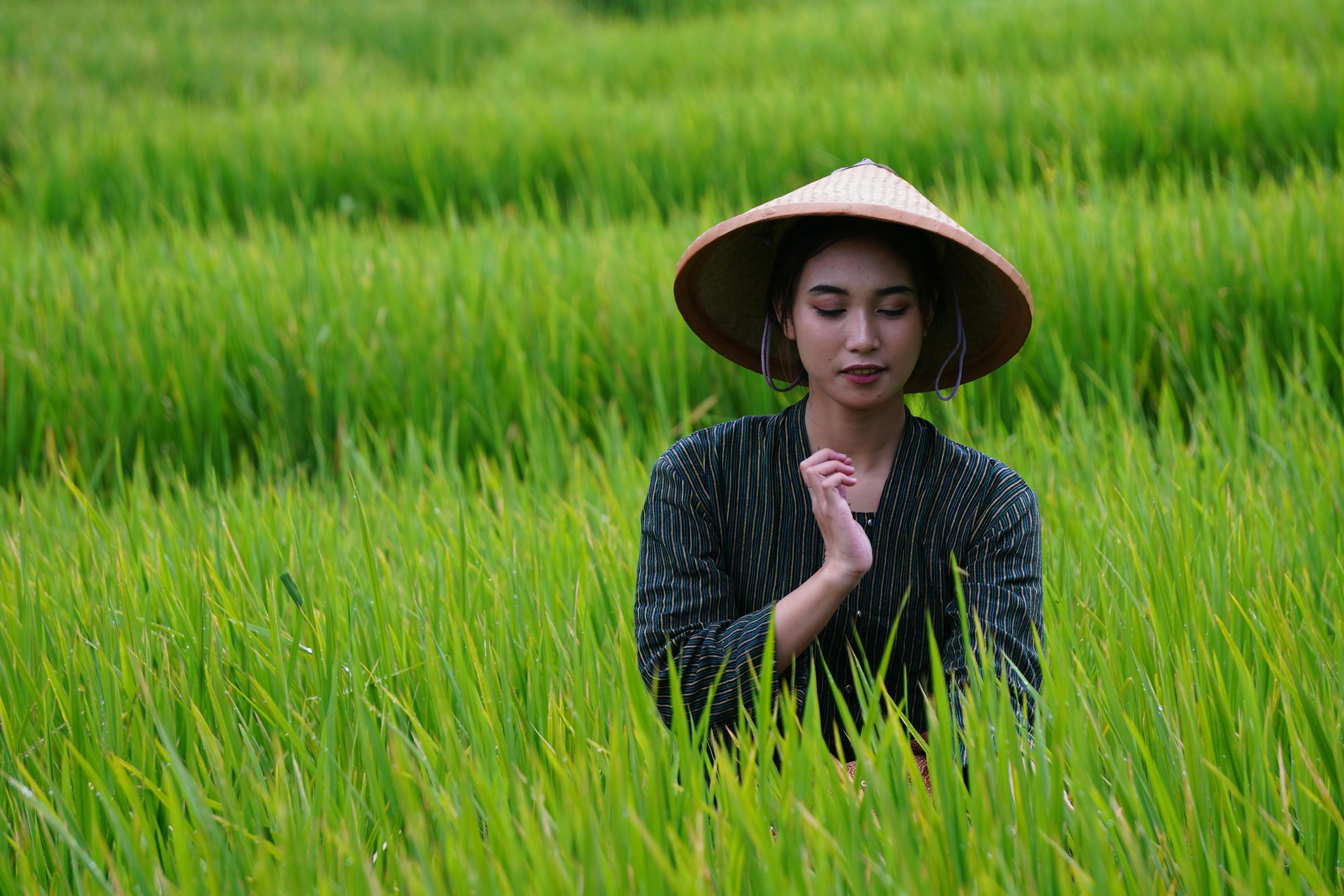An Asian Woman in a Straw Hat sitting peacefully in a grassy field.