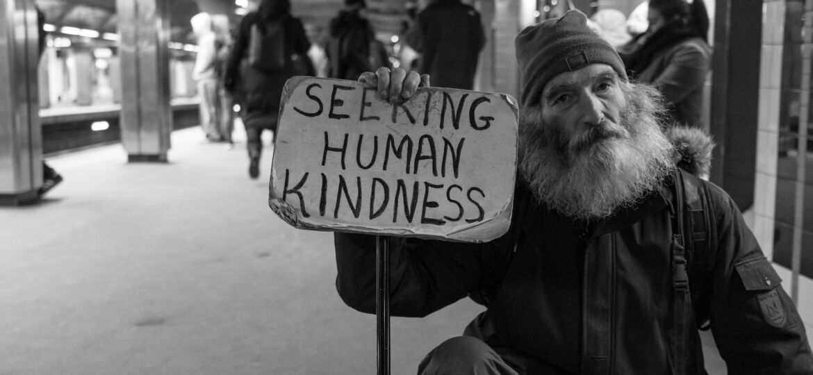 A man with a greying beard holding a sign that says "seeking human kindness"