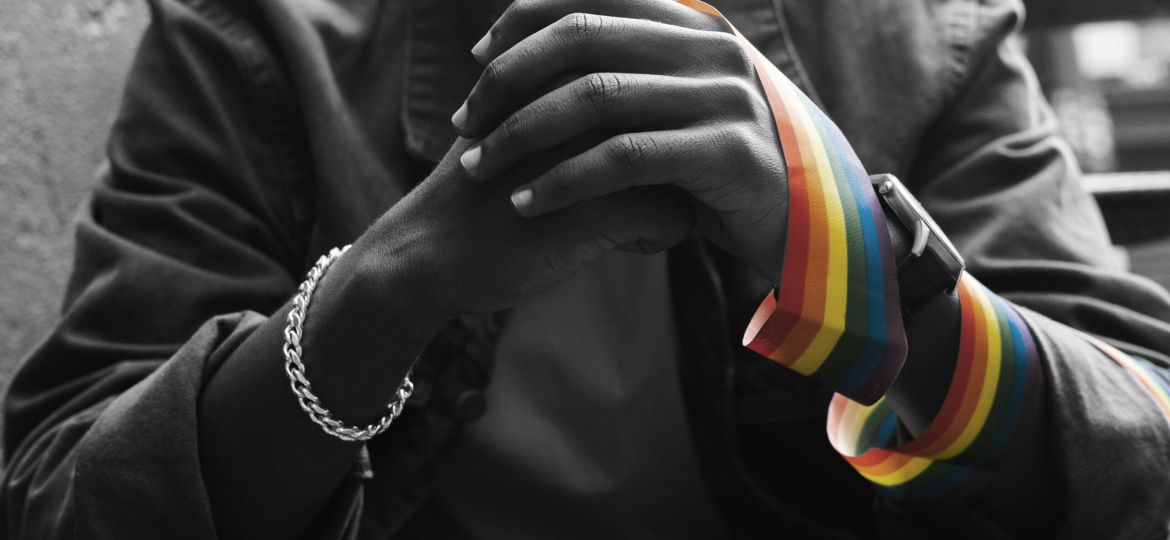 A cropped picture of a black man holding his hands with a rainbow pride ribbon wrapped around one wrist
