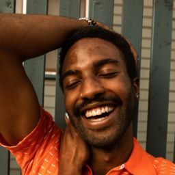 A black man wearing an orange shirt smiling brightly